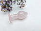 A pale pink rose quartz butt plug with a smooth, polished finish, resting on a white textured surface with dried purple flowers in the background.