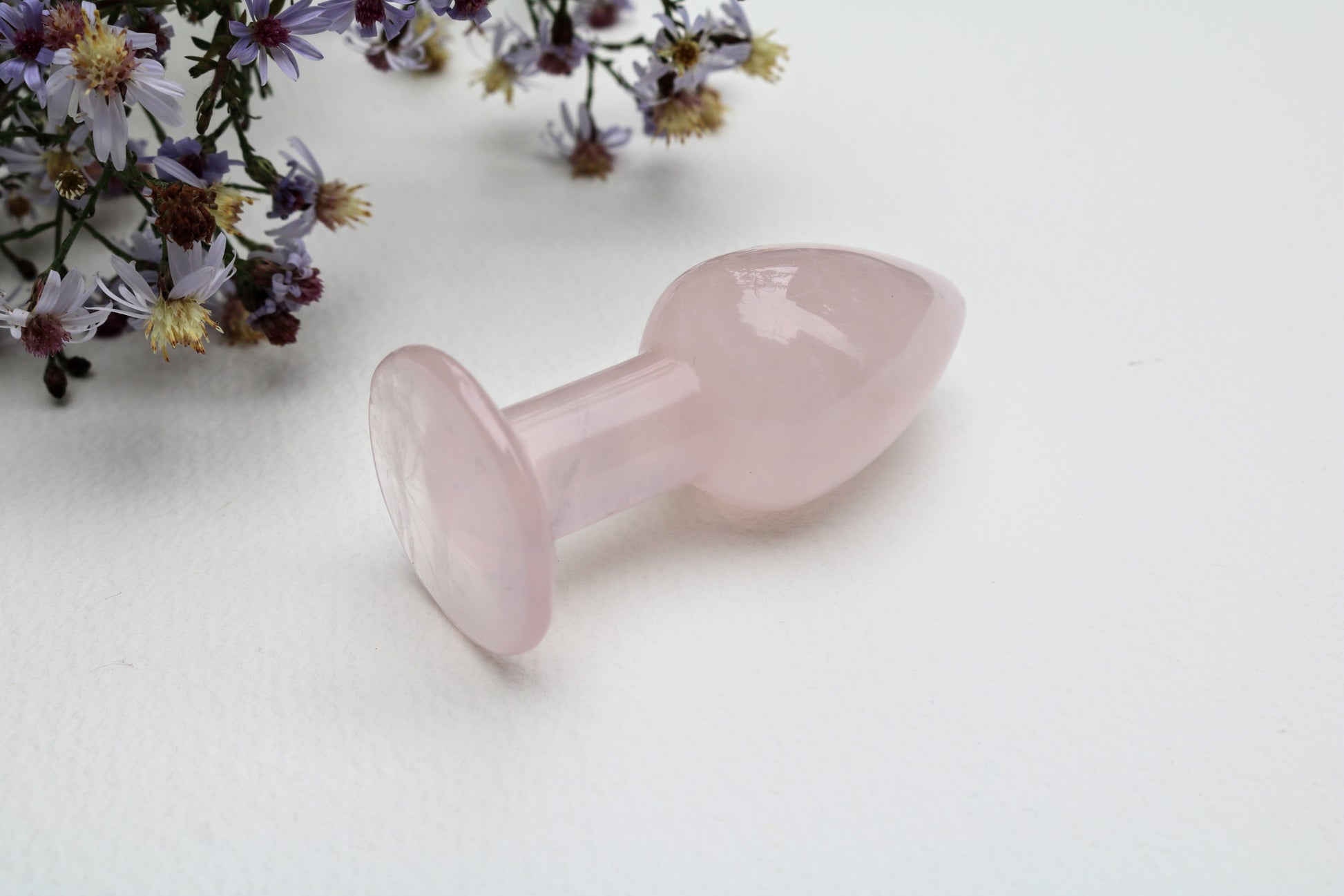 A translucent light pink rose quartz butt plug with a flared base, displayed on a clean white surface with wildflowers in the upper left corner.