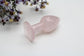 A translucent light pink rose quartz butt plug with a flared base, displayed on a clean white surface with wildflowers in the upper left corner.