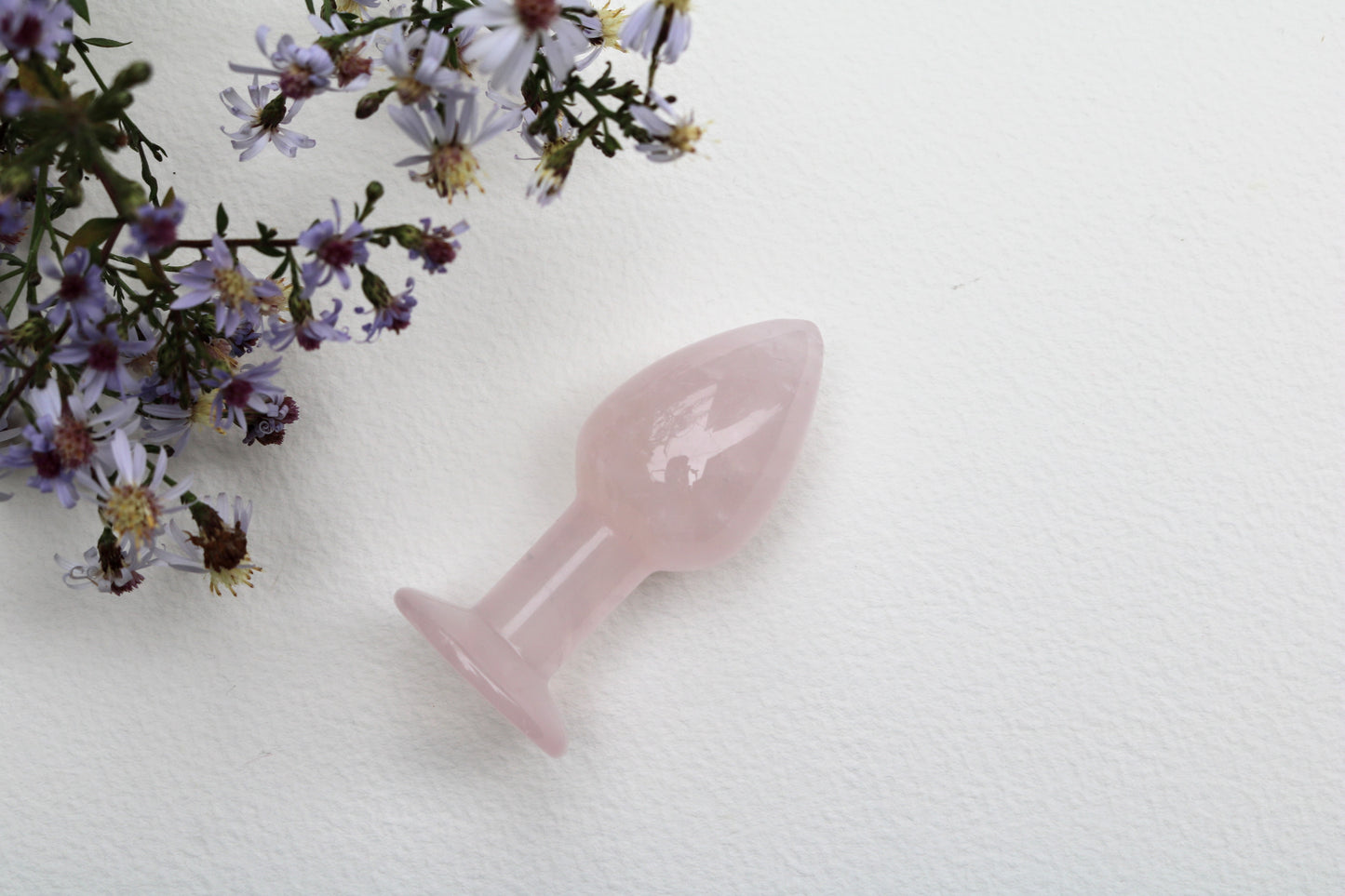 Small, polished rose quartz butt plug with a tapered tip and flat base, lying on a white textured surface with a  wildflowers on the corner