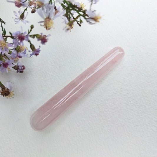 A polished pink rose quartz crystal wand lies diagonally on a textured white surface, styled with delicate purple and white wildflowers in the upper left corner.