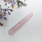 A polished pink rose quartz crystal wand lies diagonally on a textured white surface, styled with delicate purple and white wildflowers in the upper left corner.