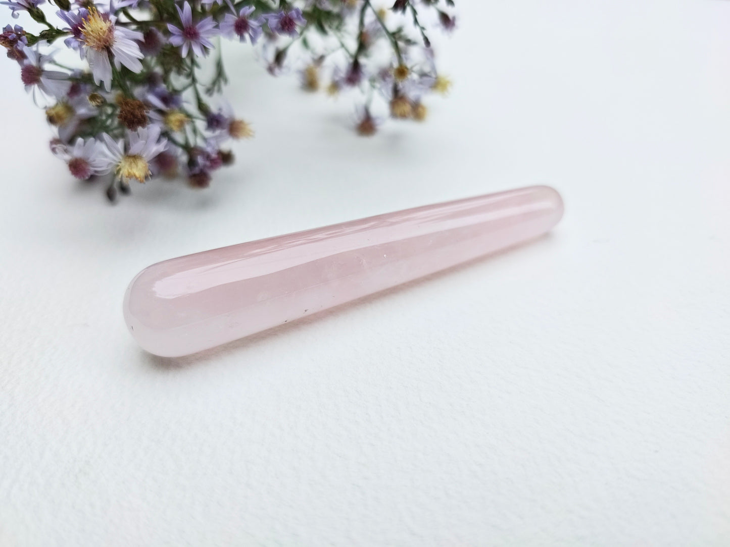 A polished pink rose quartz crystal point resting on a white textured surface, with small wildflowers in the upper left corner.