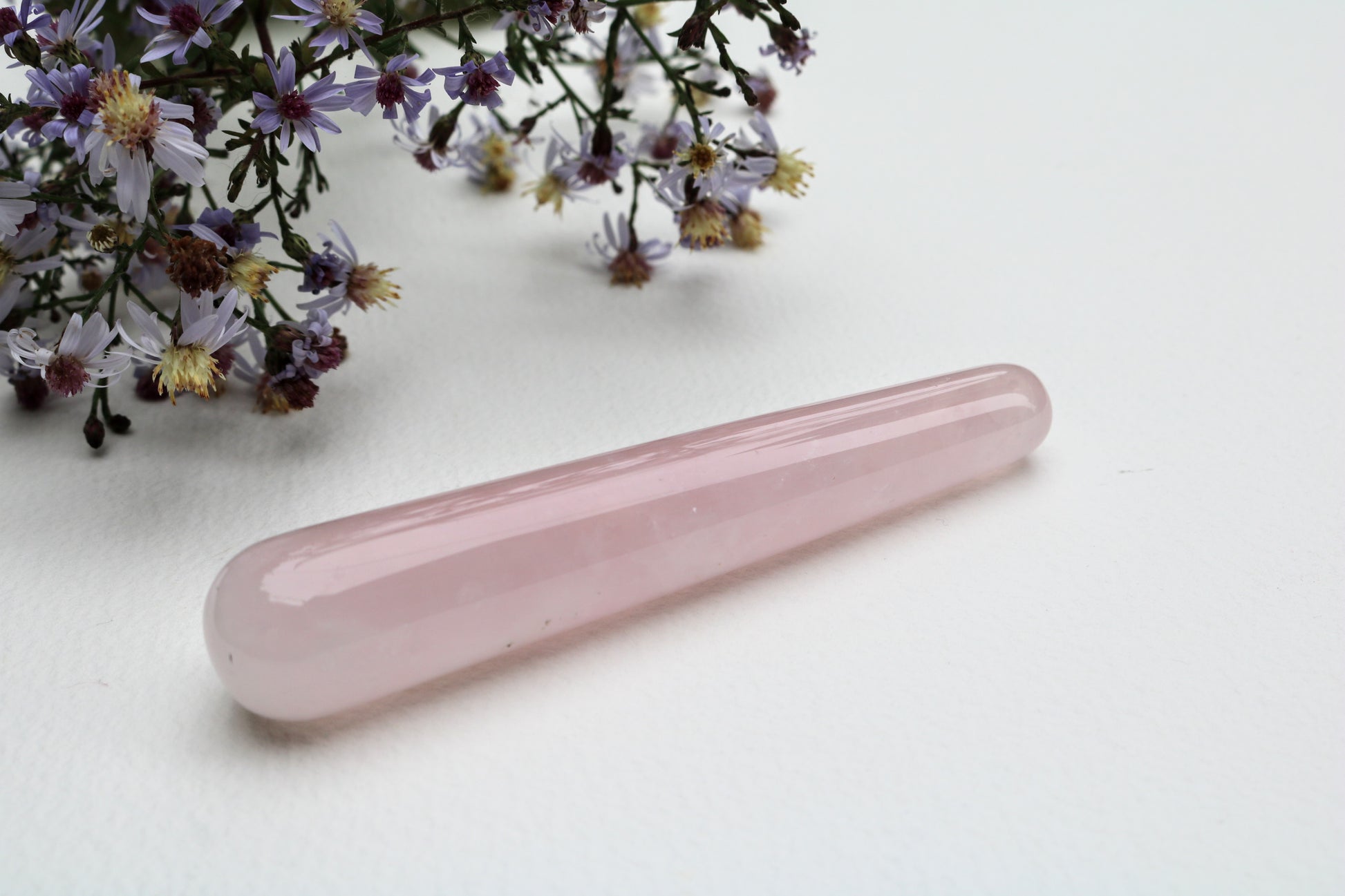 A polished pink rose quartz crystal wand lies on a white surface next to wildflowers.