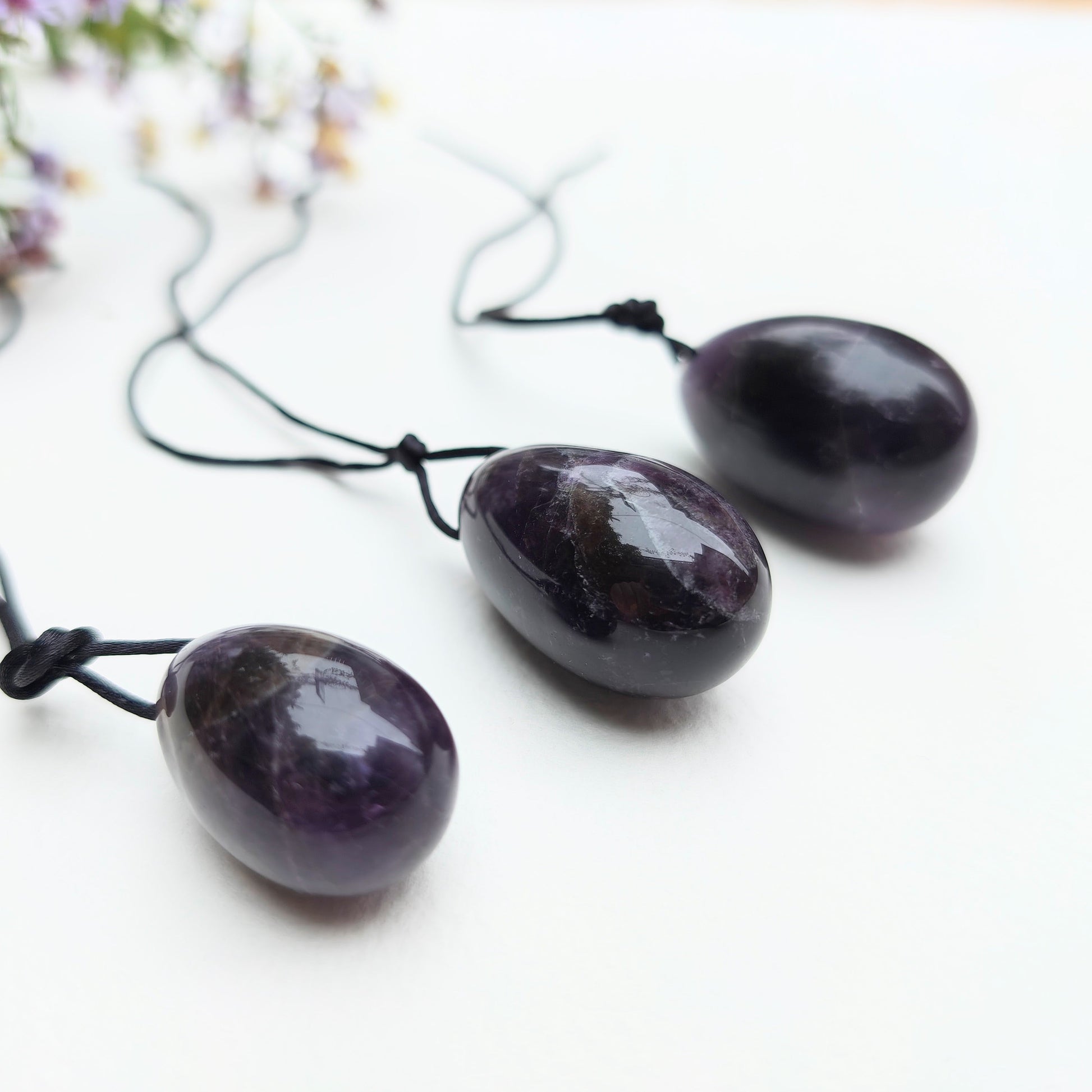 A set of three polished Amethyst yoni eggs in varying shades of purple, with black cords, lying on a white textured surface with wildflowers.