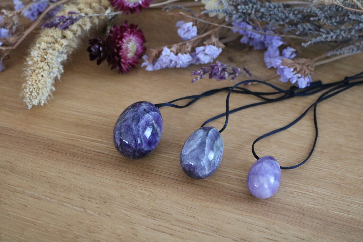 A set of three polished Amethyst yoni eggs in varying shades of purple, with black cords, lying on a wooden surface with dried flowers.