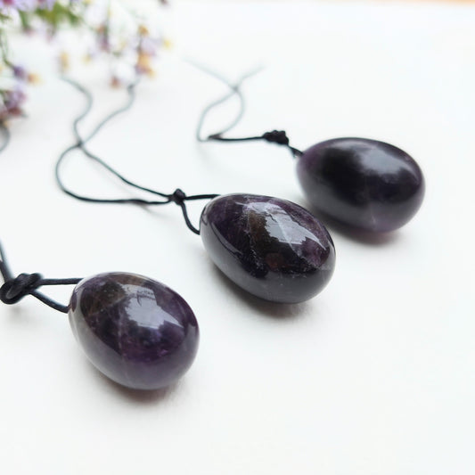 A set of three polished Amethyst yoni eggs in varying shades of purple, with black cords, lying on a white textured surface with wildflowers.