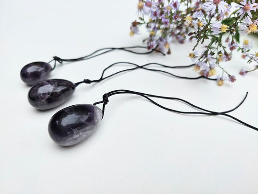 A set of three polished Amethyst yoni eggs, with black cords, lying on a white textured surface with wildflowers.