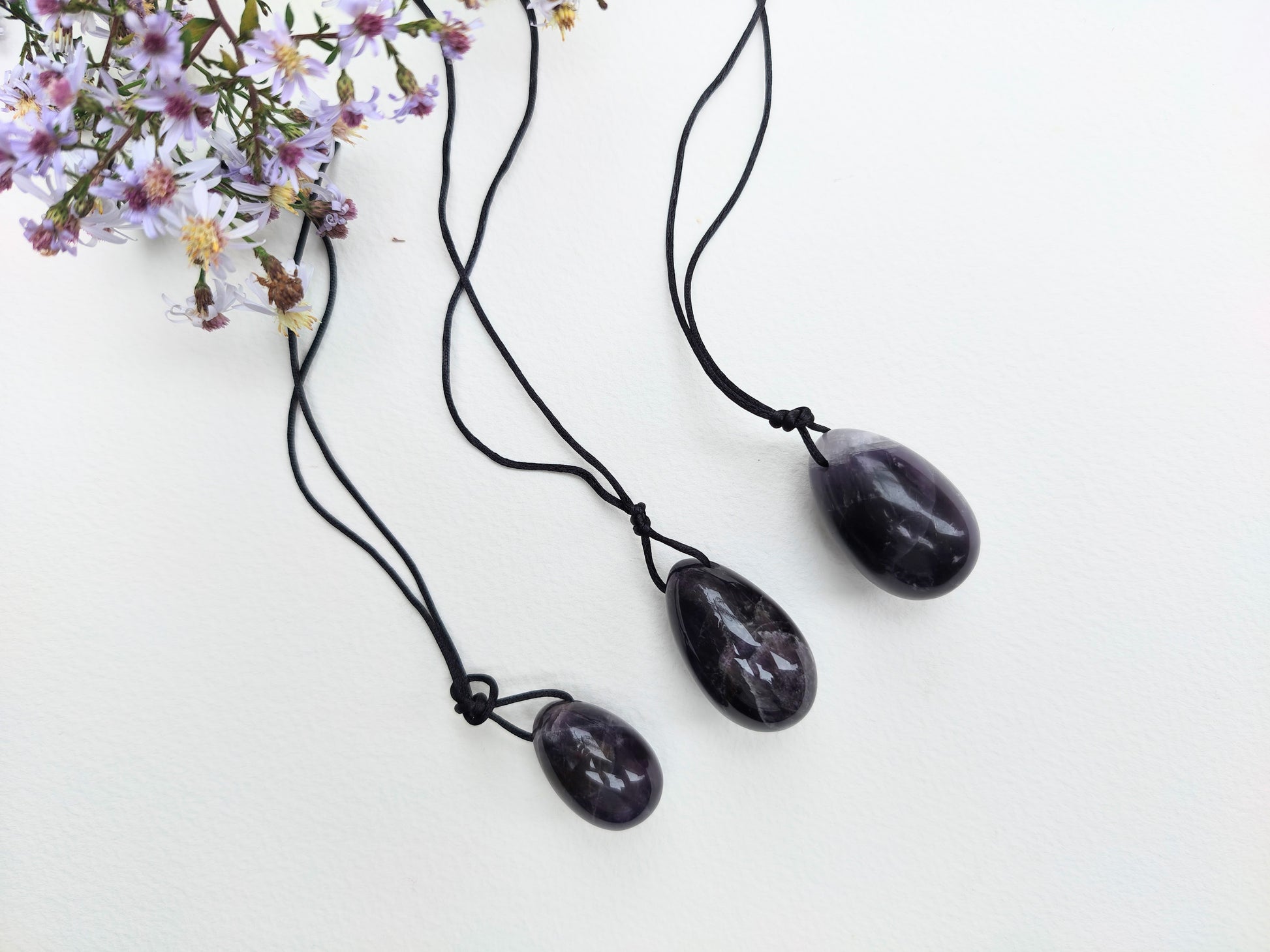 A set of three polished Amethyst yoni eggs in varying shades of purple lying on a white textured surface with wildflowers.
