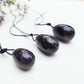 A set of three polished Amethyst yoni eggs in varying shades of purple, with black cords, lying on a white textured surface with wildflowers.