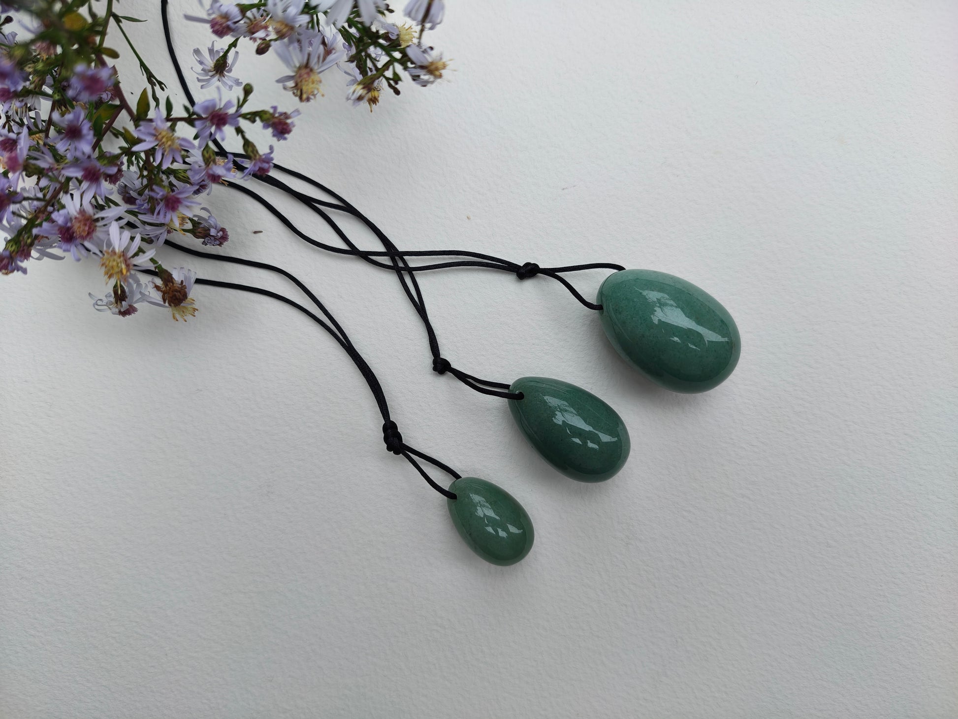 A set of three polished Green Aventurine yoni eggs, with black cords, lying on a white textured surface with wildflowers.