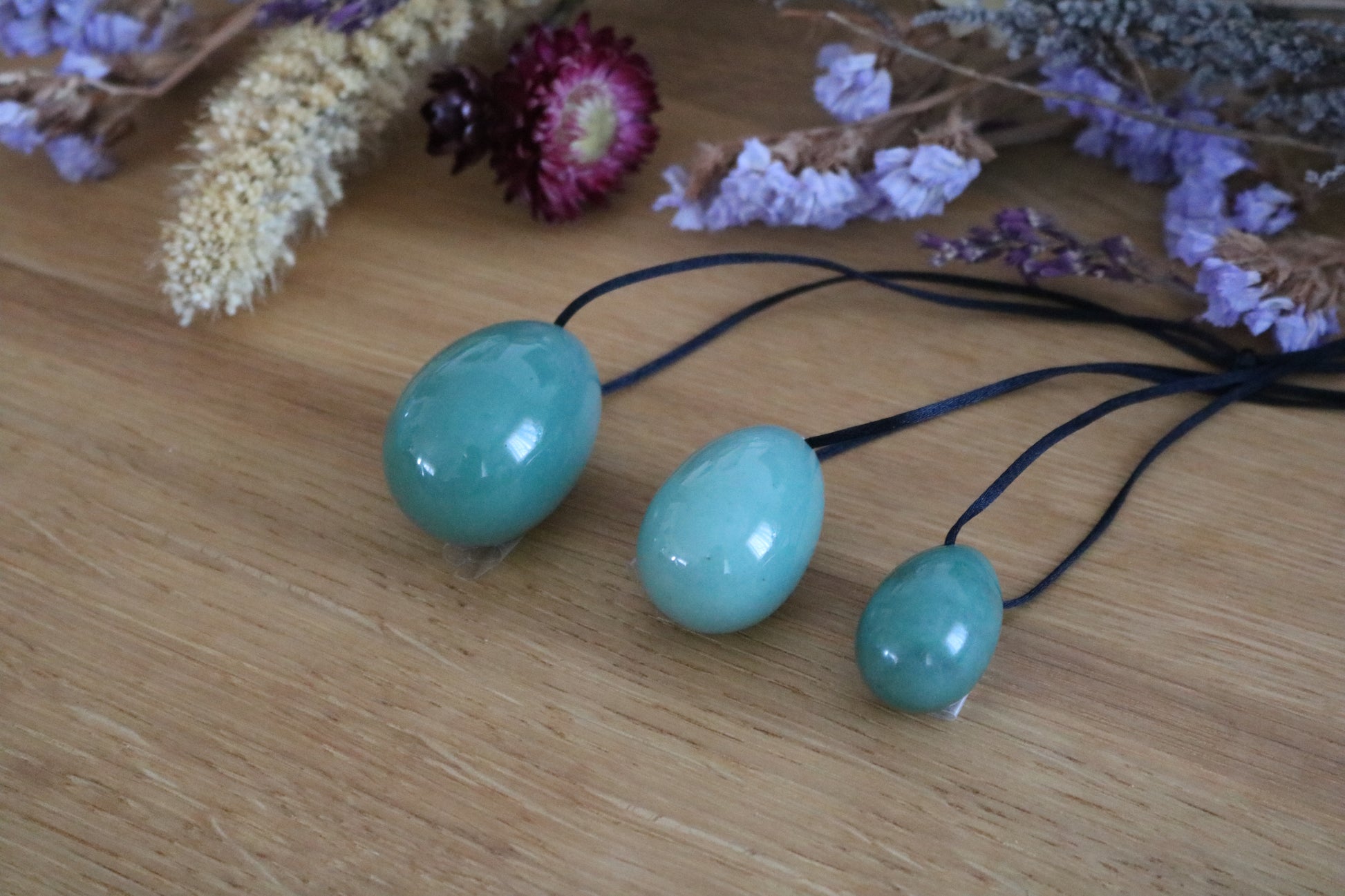 A set of three polished Green Aventurine yoni eggs in varying shades of green, with black cords, lying on a wooden surface with dried flowers.