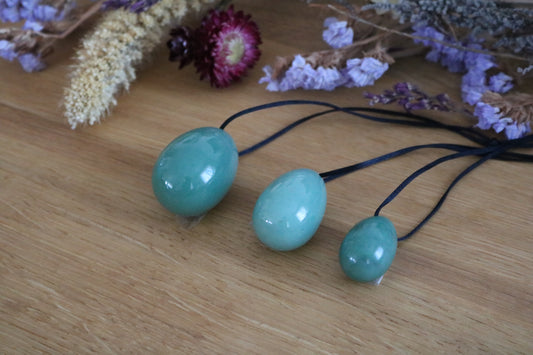 A set of three polished Green Aventurine yoni eggs in varying shades of green, with black cords, lying on a wooden surface with dried flowers.
