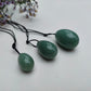A set of three polished Green Aventurine yoni eggs in varying shades of green, with black cords, lying on a white textured surface with wildflowers.