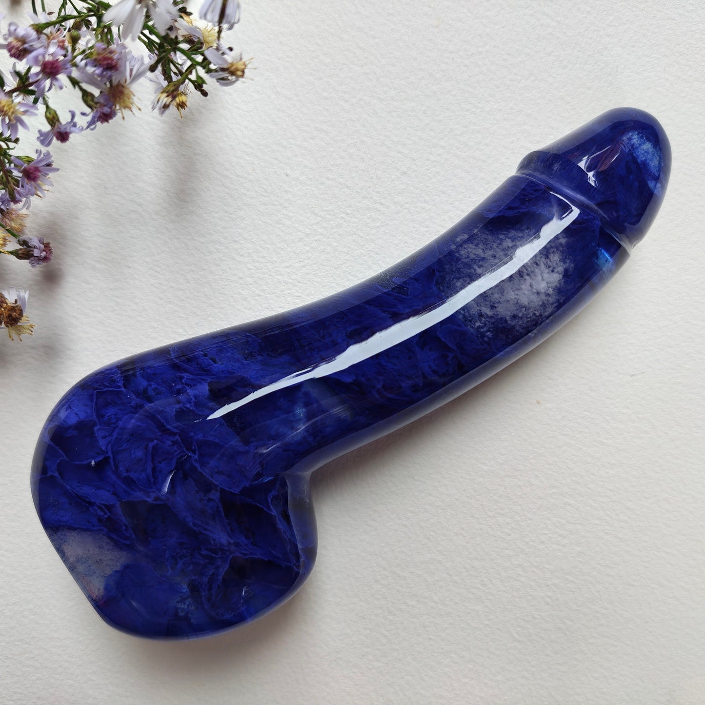 A glossy melting blue stone dildo with natural marbling, displayed diagonally on a textured off-white background with small wildflowers in the top-left corner.