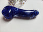 A glossy, translucent deep blue glass dildo with natural bubble and swirl patterns, resting on a white textured surface with small wildflowers in the background.