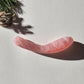 Large, curved, dual-sized rose quartz crystal wand with one thick and one thin end, lying on a light surface with pine needles and shadows.