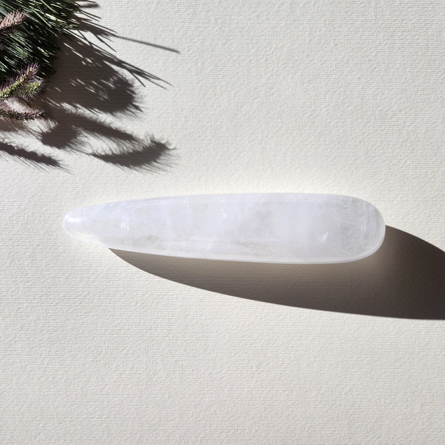 A close-up, of a large, straight, smooth Clear Quartz Yoni Wand. The lighting highlights the polished, reflective surface and the cloudy white interior with internal fractures. It is set against a light, textured background, and a sprig of dried grass. Perfect for anal stimulation