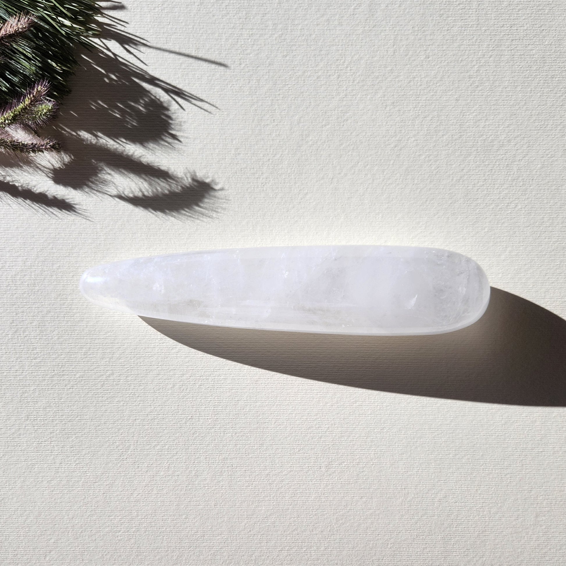 A close-up, of a large, straight, smooth Clear Quartz Yoni Wand. The lighting highlights the polished, reflective surface and the cloudy white interior with internal fractures. It is set against a light, textured background, and a sprig of dried grass. Perfect for anal stimulation