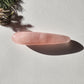Polished Rose Quartz Yoni Wand (large, straight) with a soft pink hue and natural internal striations. The crystal rests on a light-colored background, with a sprig of pine needles casting a shadow. Perfect for sexual stimulation