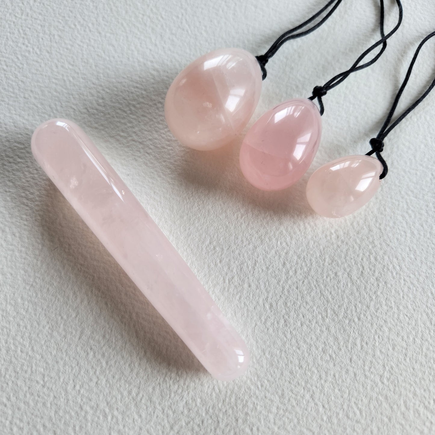 A set of three polished rose quartz yoni eggs with black strings and a smooth rose quartz mini wand, lying on a white textured surface.
