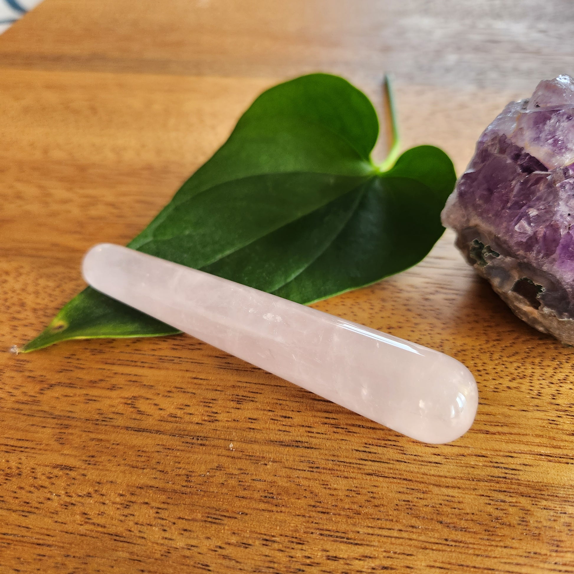 Small, smooth pink rose quartz crystal wand with a rounded end, placed on a wooden surface next to a green leaf and a rough amethyst geode.