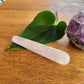 Small, smooth pink rose quartz crystal wand with a rounded end, placed on a wooden surface next to a green leaf and a rough amethyst geode.