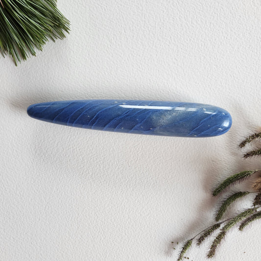 Polished blue aventurine crystal yoni wand with subtle shimmer and a smooth, tapered form for sex anal to reach the G-Spot, displayed on a white backdrop with pine sprigs and dried foliage. 
