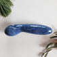 Large, polished blue aventurine crystal wand with a unique contoured, spoon-like shape, lying on a white surface with pine needles and dried foliage