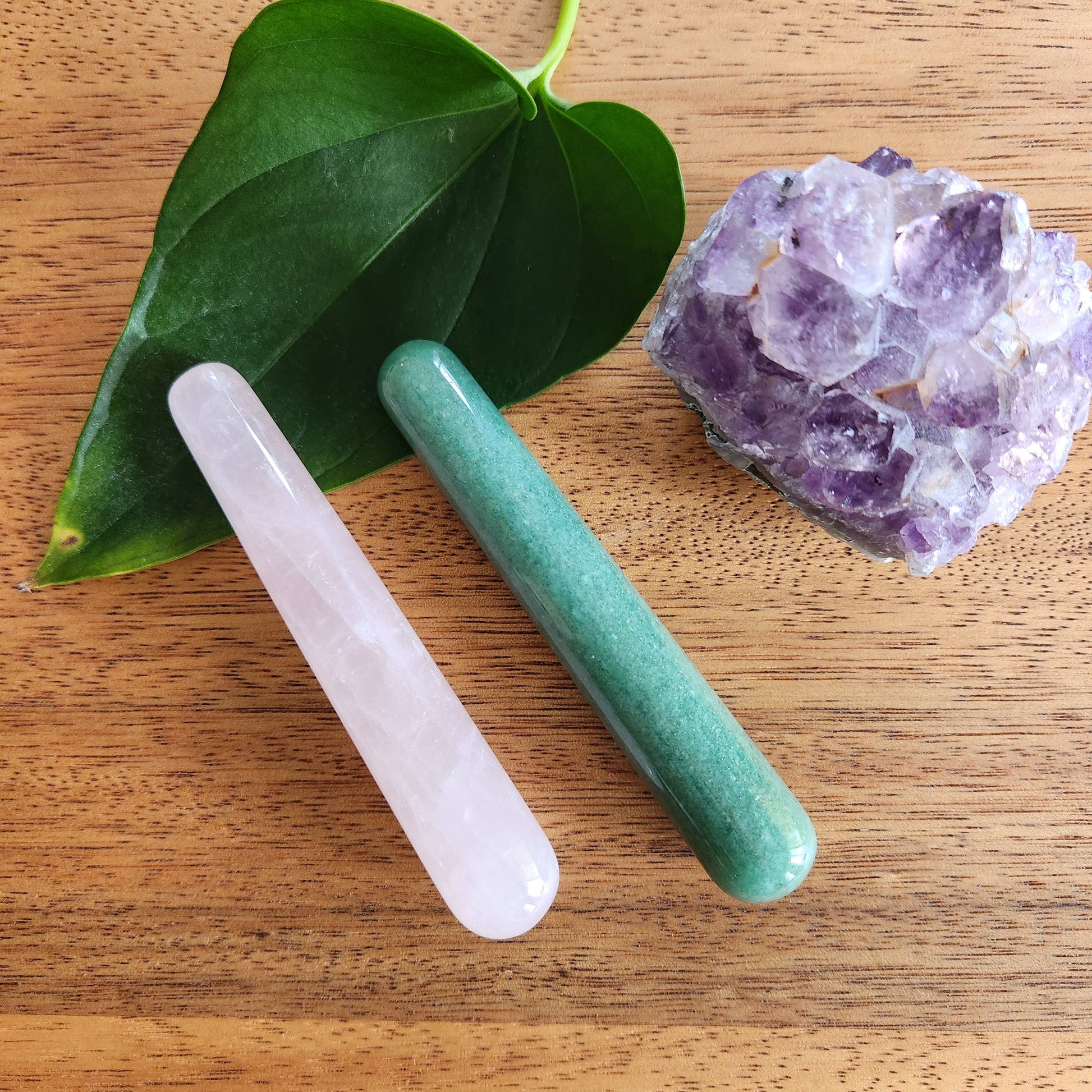 Two mini crystal wand in Rose quartz and Green aventurine on a wood surface with a Crystal rock and a green leaf 