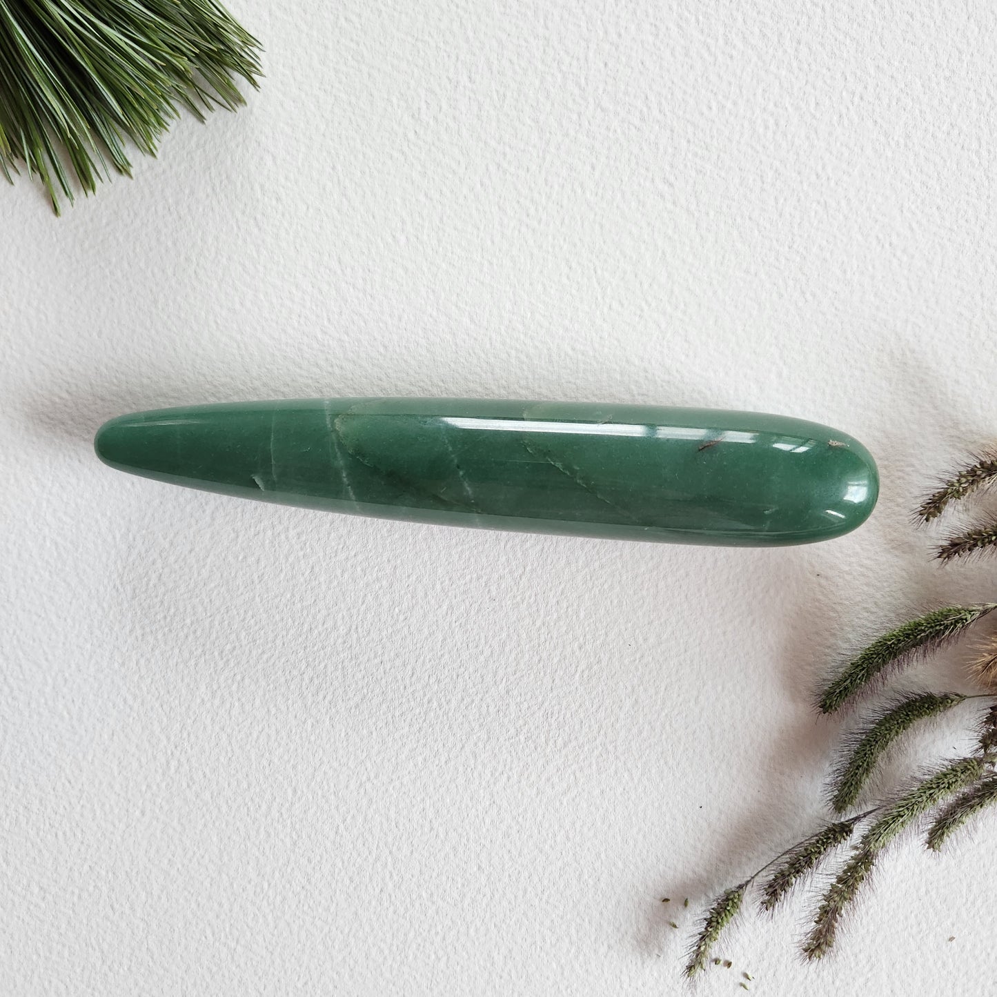Polished green aventurine crystal yoni wand with a smooth, tapered shape to reach the G-Spot, shown on a white background with pine needles and dried grass.