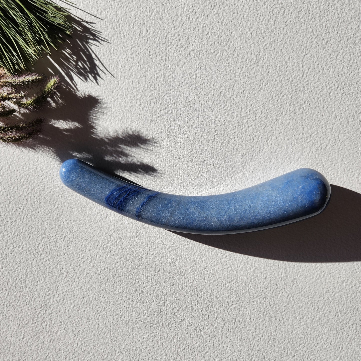 Smooth, crescent-shaped blue aventurine crystal wand with a uniform thickness, lying on a light surface with pine needles and shadows from a plant.