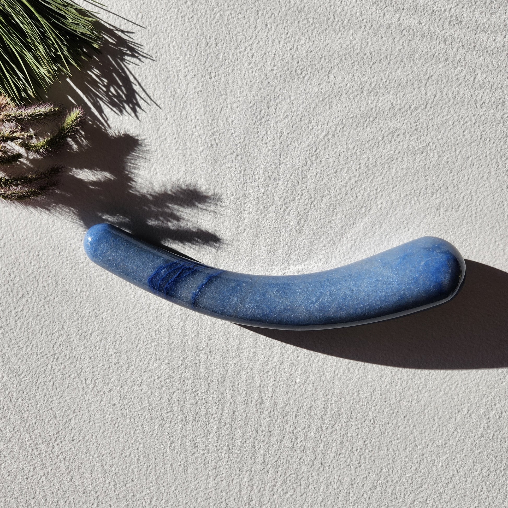 Smooth, crescent-shaped blue aventurine crystal wand with a uniform thickness, lying on a light surface with pine needles and shadows from a plant.