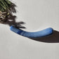 Smooth, crescent-shaped blue aventurine crystal wand with a uniform thickness, lying on a light surface with pine needles and shadows from a plant.