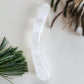 Smooth, crescent-shaped clear quartz crystal wand with a uniform thickness, lying on a light surface with pine needles and dried foliage.