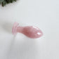 A hand-polished, curved rose quartz Butt-Plug lying on a light-colored surface with a green pine branch in the background.