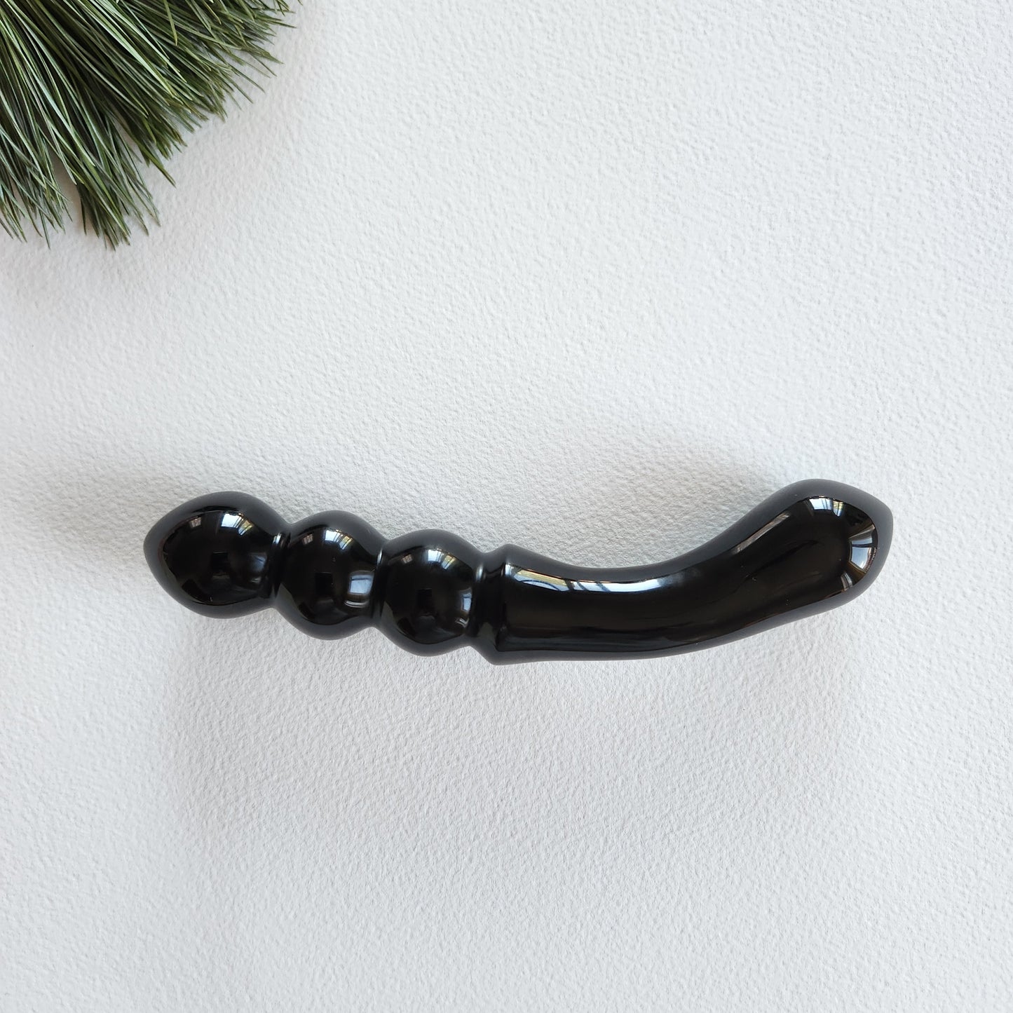Smooth, double-ended black obsidian crystal wand with a gently curved end and a beaded or segmented end, lying on a white surface with pine needles.