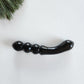 Smooth, double-ended black obsidian crystal wand with a gently curved end and a beaded or segmented end, lying on a white surface with pine needles.