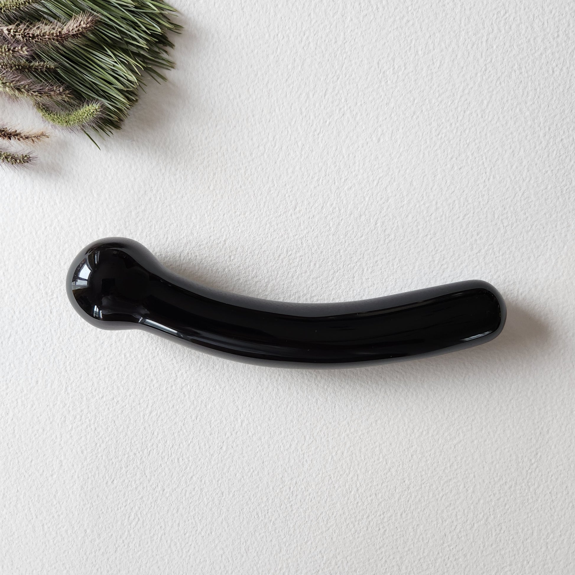 A hand-polished, curved black obsidian crystal wand with round tip lying on a light-colored surface with a green pine branch in the corner.