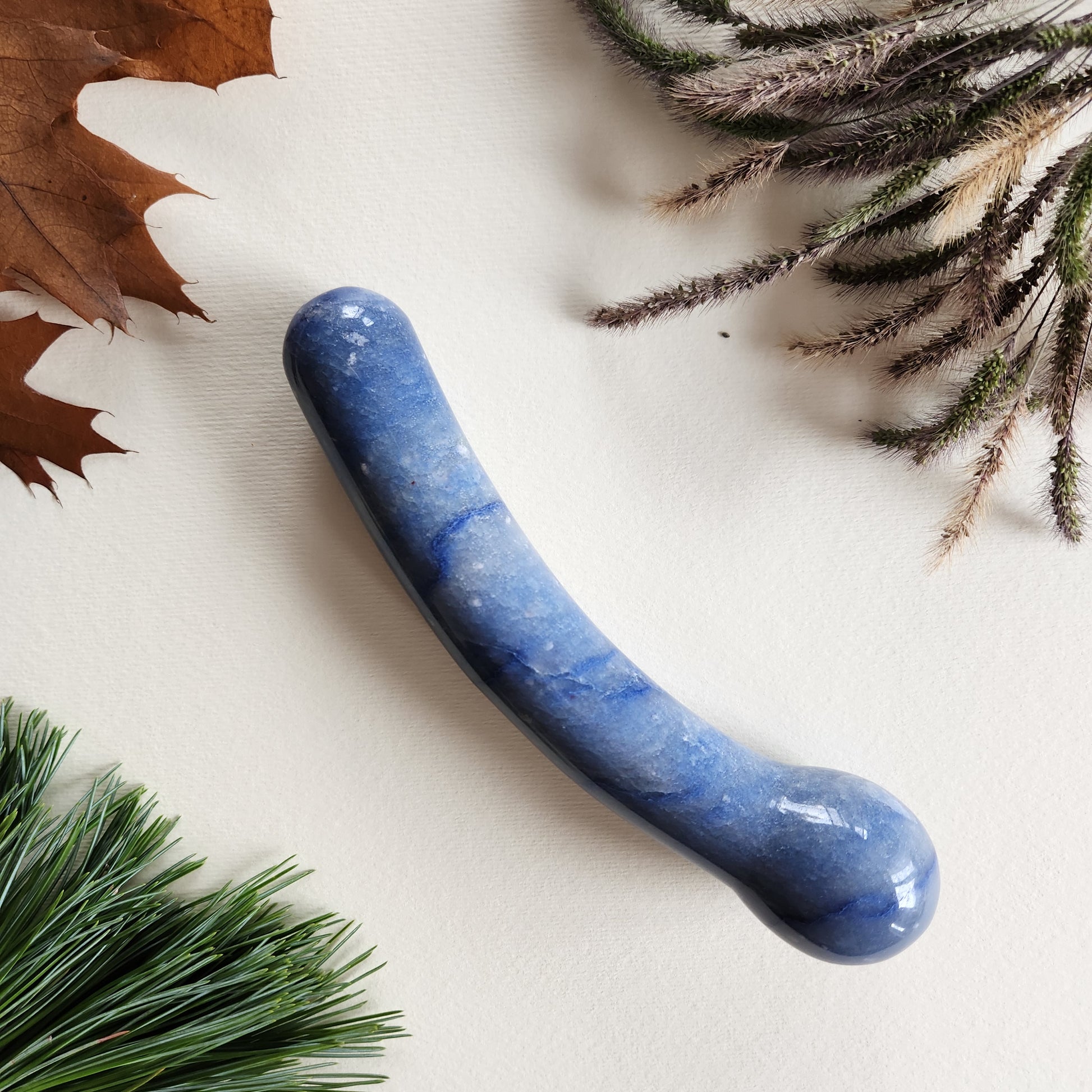 Curved blue aventurine crystal wand with a smooth, polished finish, designed for G-spot stimulation, lying on a light surface surrounded by autumn leaves and dried foliage.