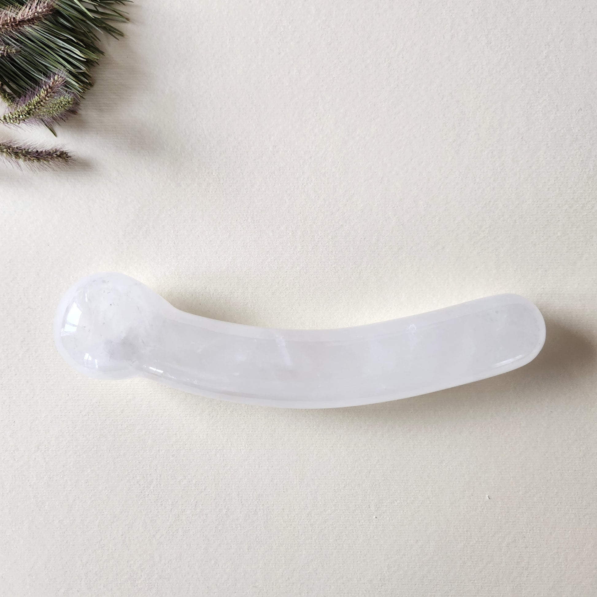 Curved clear quartz crystal wand with a smooth, polished finish, designed for G-spot stimulation, lying on a light surface with pine needles and dried foliage.