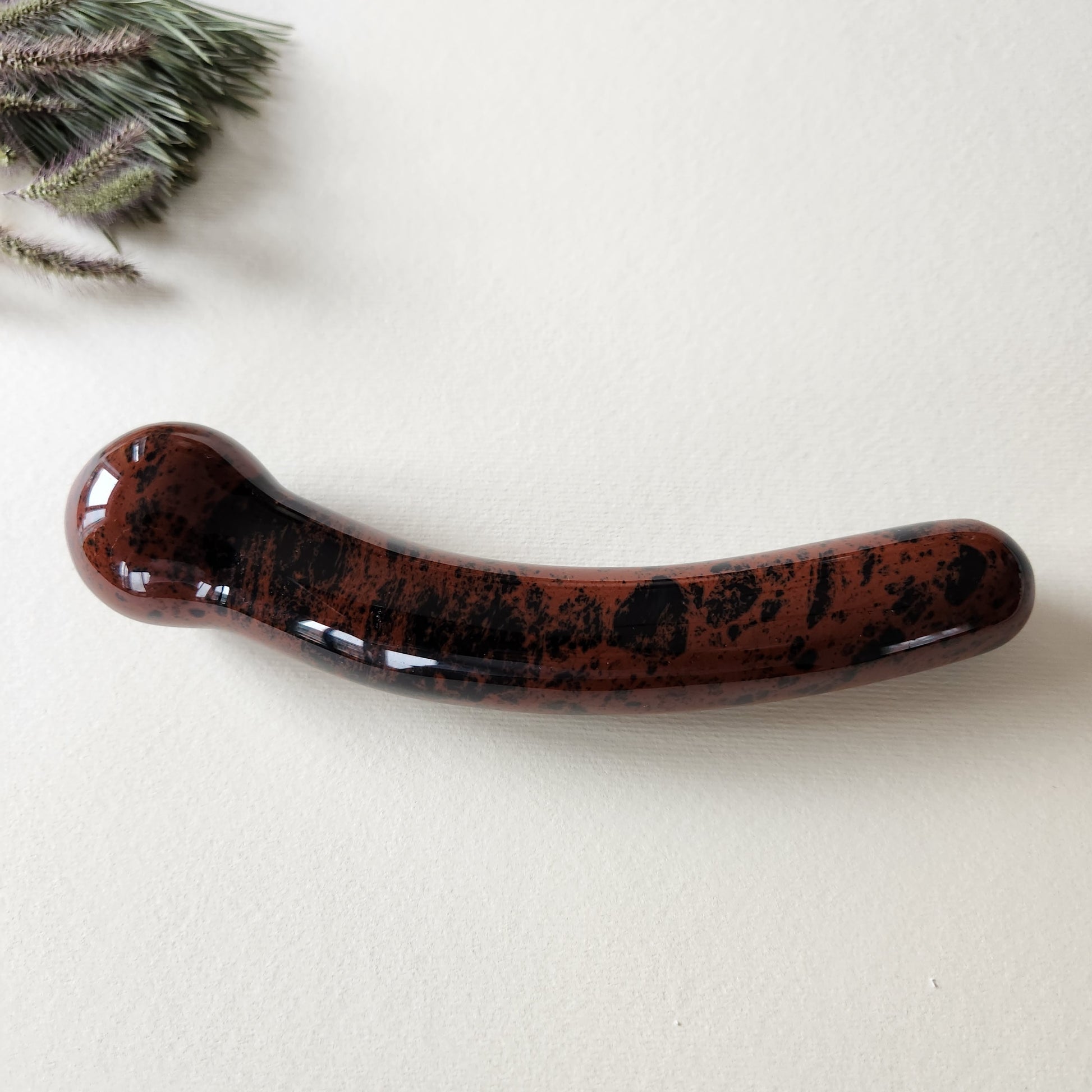 Curved mahogany obsidian crystal wand with a smooth, polished finish in reddish-brown with black markings, designed for G-spot stimulation, lying on a light surface with pine needles and dried foliage.