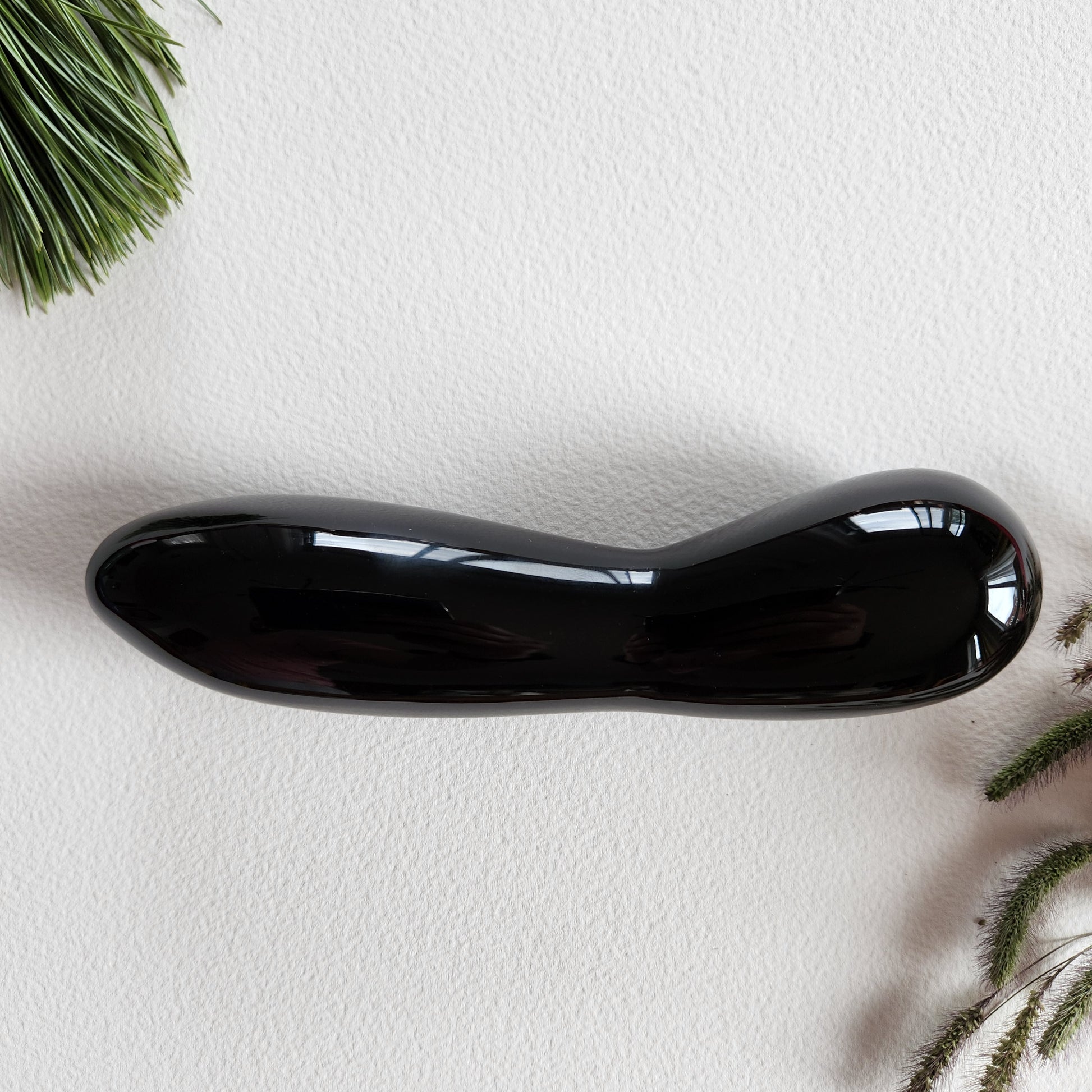 Large, polished black obsidian crystal wand with a unique contoured, spoon-like shape, lying on a white surface with pine needles and dried foliage.