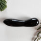 Large, polished black obsidian crystal wand with a unique contoured, spoon-like shape, lying on a white surface with pine needles and dried foliage.