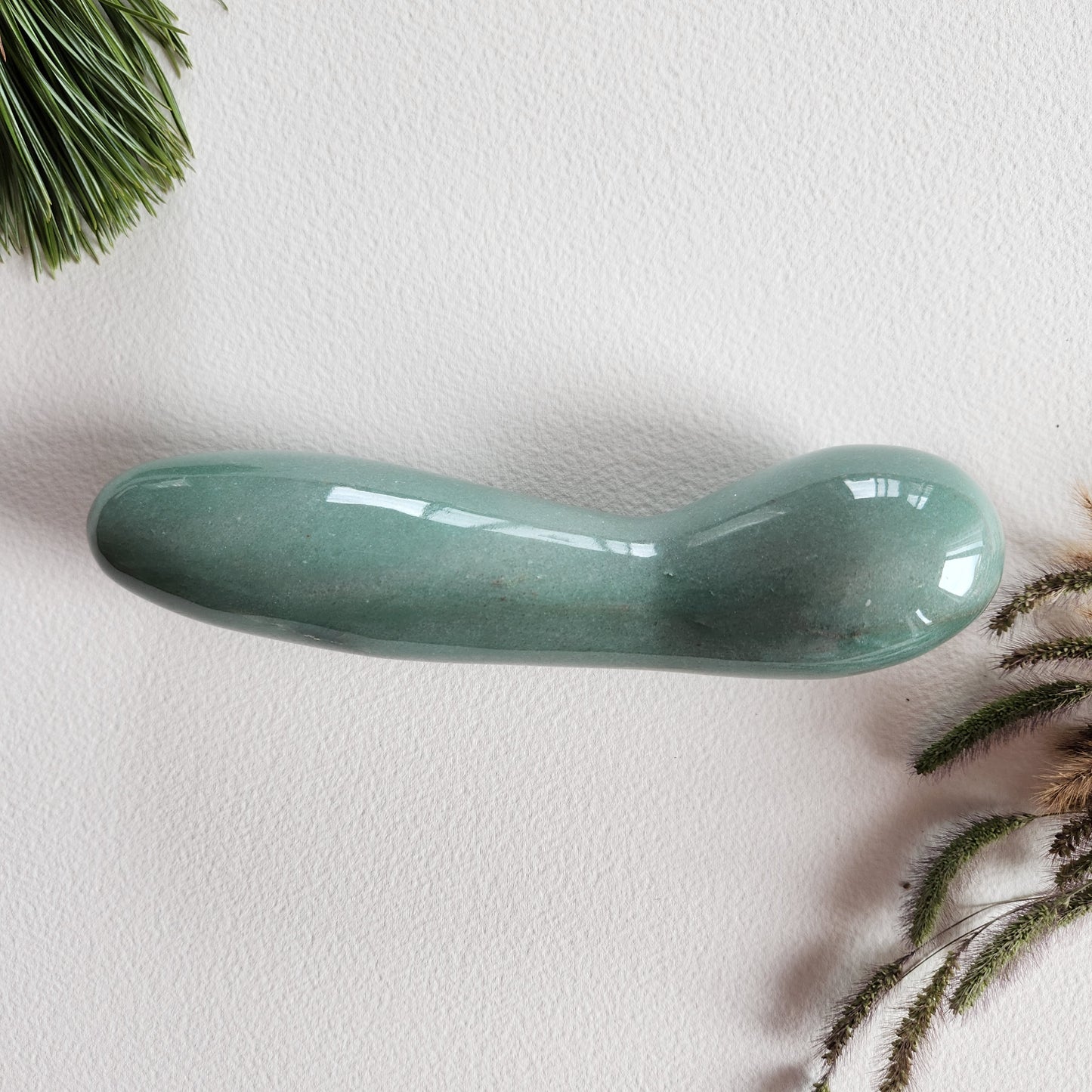 Large, polished green aventurine crystal wand with a unique contoured, spoon-like shape, lying on a white surface with pine needles and dried foliage.