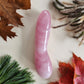 Large, curved, rose quartz crystal yoni wand with a smooth, polished finish, lying on a light surface surrounded by colorful autumn leaves and pine needles.