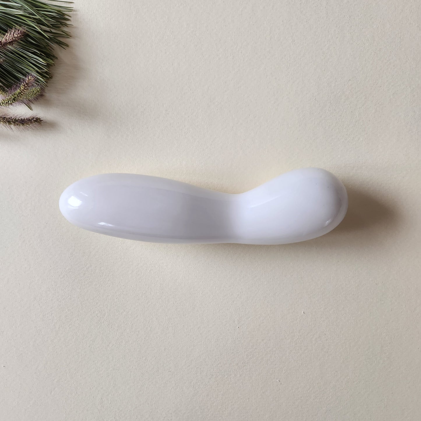 Large, polished white jade crystal wand with a unique contoured, spoon-like shape, lying on a light surface with pine needles and dried foliage.