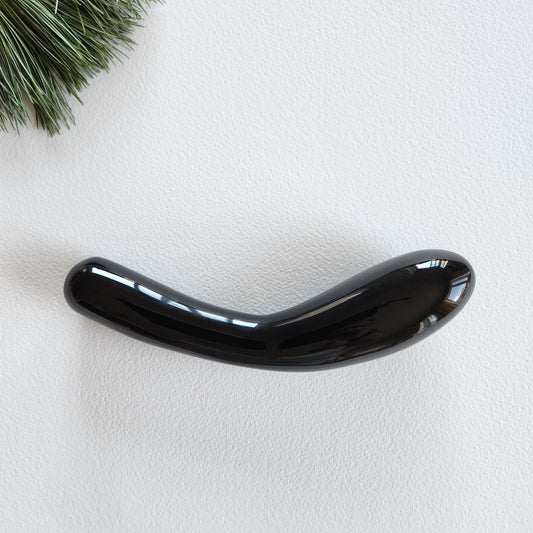 Large, curved, dual-sized black obsidian crystal wand with one thick and one thin end, lying on a light surface with pine needles.