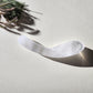Large, curved, dual-sized clear quartz crystal wand with one thick and one thin end, lying on a light surface with pine needles and dried foliage.