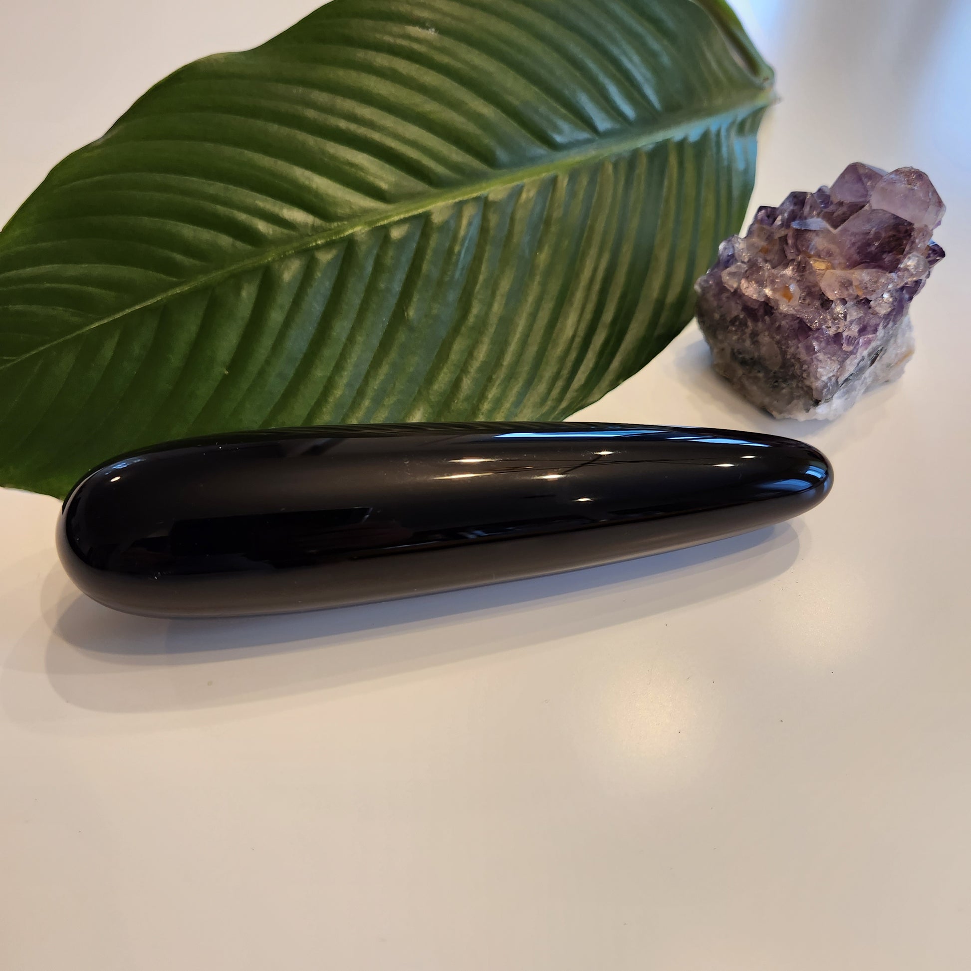 Thick, polished black obsidian crystal wand with a large diameter to reach the G-Spot, lying on a white surface next to a large green leaf and a rough amethyst geode.